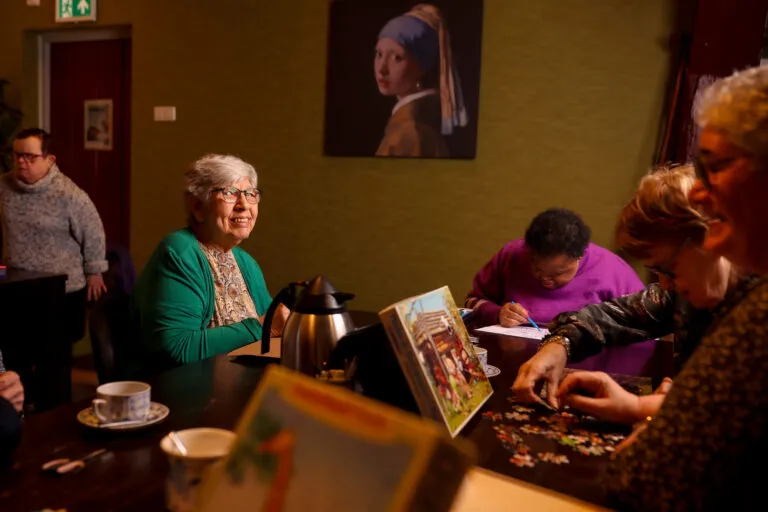Doelgroep ouderen, cliënt lacht in camera en zit aan tafel waar wordt gepuzzeld en getekend