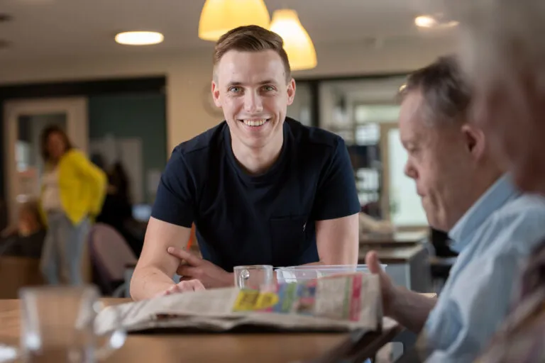 Amarant als werkgever, zorgmedewerker zit aan tafel met cliënten en kijkt en lacht in de camera