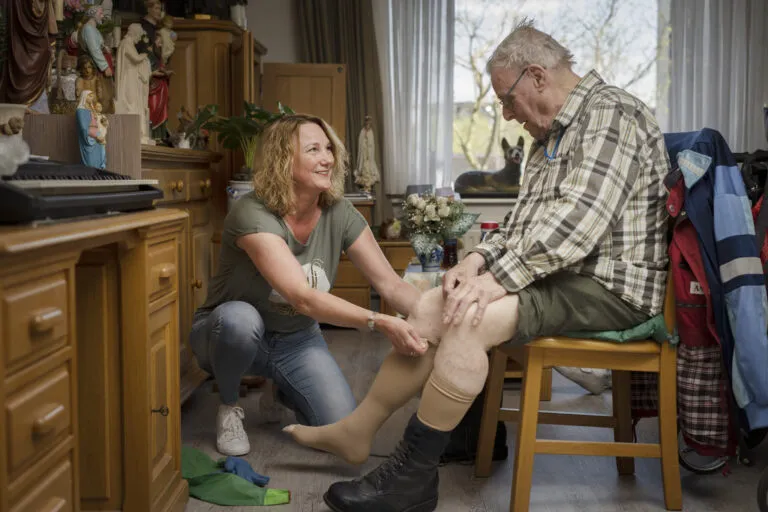 Doelgroep ouderen, zorgprofessional trekt steunkousen bij cliënt aan.