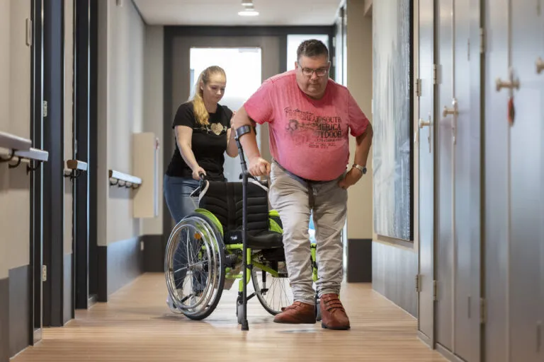 Doelgroep NAH, Niet-Aangeboren Hersenafwijking. Zorgprofessional staat met rolstoel en cliënt staat op uit rolstoel met kruk.